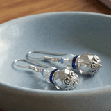A pair of silver-toned dangle crystal earrings with faceted clear beads, small blue accents, and filagree details, resting on a light blue ceramic dish - Lapanda Designs - Parade Handmade