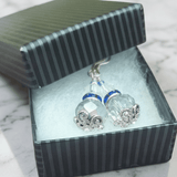 A pair of silver-toned dangle earrings with faceted clear grey and blue crystal beads with royal blue crystal band accents, presented in an open striped gift box on a light marble surface - Lapanda Designs - Parade Handmade