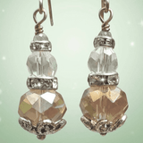 A pair of handmade dangle earrings featuring faceted clear beads with a soft iridescent topaz and gold tint, paired with silver metal beadcaps and sterling silver hooks, on a light green sparkly background - Lapanda Designs at Parade Handmade