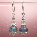 A pair of silver-tone dangle earrings with faceted clear crystal beads and large iridescent blue-grey crystal drops, displayed against a soft pink background