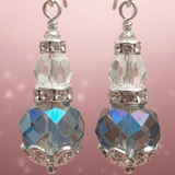 A pair of dangling earrings with silver alloy details and clear faceted crystal beads, each hanging from a Sterling silver hook. The lower beads are large iridescent blue and grey beads with silver filigree detailing at the bottom, set against a soft pink background. By Lapanda Designs - Parade Handmade