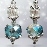 A pair of dangling crystal earrings with large faceted iridescent blue-green beads, silver-toned ornate caps, topped with smaller clear cut crystal beads above, shown on a soft light gray background with subtle sparkles - Lapanda Designs - Parade Handmade