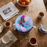 A light blue knitted tea cosy pot sock with colorful feathers and flower applique, on a teapot on a rustic wooden table surrounded by teacups, a newspaper, and oranges.