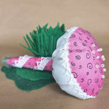 A handmade fabric mushroom or decorative pincushion with a pink patterned cap, white ruffled trim, green felt stand, and a pink wrapped handle, displayed on a tan surface.
