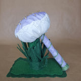 A handmade felt mushroom decoration or ornament with a pale lavender cap, white lace trim, seed bead accents, a lavender stem wrapped in green cord, and a dark green felt base of grass, photographed against a plain tan backdrop.