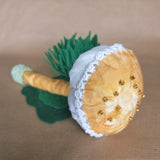 A handmade mushroom-shaped pincushion with a mustard cotton cap decorated with white lace trim and small gold beads, a mustard stem, green felt leaves, displayed on a brown cardboard background.