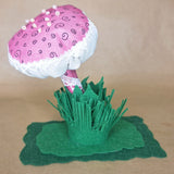 A cotton mushroom with a raspberry pink head and stem decorated with lace trim and seed beads all on a green felt grass base photographed against a tan background.