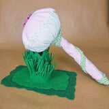 A handmade fabric mushroom ornament or pincushion with a pink and white striped head, green lace trim, a striped stem, and a green felt grass base, displayed on a tan background.