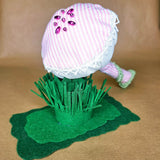 A handmade mushroom keepsake with a pink and white striped fabric cap, white lace trim, and pink stem, sitting on green felt grass and a green felt base against a tan background.