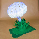 A handmade fabric mushroom decoration with a pale lavender cap adorned with small seed beads and white lace trim, set on a purple fabric stem on a green felt base with green felt grass, photographed against a plain tan background.