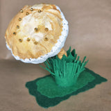A miniature fabric mushroom figurine with a mustard and cream cap, white lace trim, small gold beads, a mustard felt stem and grass, and a green felt base, set against a plain brown background.