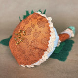 A handmade mushroom-shaped fabric ornament or decorative item, featuring an orange patterned cap with cream lace trim, a light blue underside, green felt accents, and a small cluster of gold seed beads on top, photographed on a neutral beige surface.
