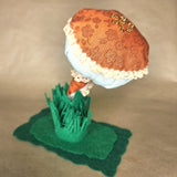 A handmade mushroom-shaped fabric ornament accessory, featuring an orange patterned cap with white lace trim, a pale blue underside, a small orange-and-white striped stem, green felt grass, and a dark green felt base, photographed against a neutral beige background.