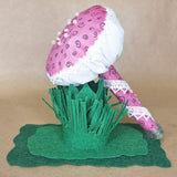 A handmade mushroom-shaped decorative object with a pink and white patterned fabric cap, green felt grass-like base, and a pink wrapped stem, displayed against a plain tan background.