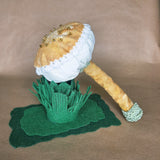 A handmade mushroom-shaped pin cushion or decorative object displayed upright on a layered green felt base against a tan paper backdrop. The top is mustard with lace trim and covered in small gold beads, while the stem is also mustard and wrapped with pale green cord at the base.