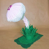 A handmade mushroom-shaped decorative object made from light pink and white striped fabric, with white lace trim, small purple beads, and green lace edging, mounted on a green felt grassy base on a tan background.