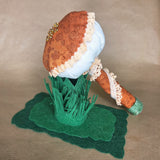 A handmade mushroom-shaped keepsake or decorative object with an orange patterned fabric cap, cream lace trim, a burmt orange fabric stem, green felt grass, and a green felt base, photographed on a beige fabric background.