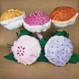 Five fabric mushroom-shaped ornaments, decorations or pincushions arranged on green felt leaves against a tan background. The top row has three larger mushrooms with white lace trim, one pink with tiny black swirls and one orange-brown floral pattern.